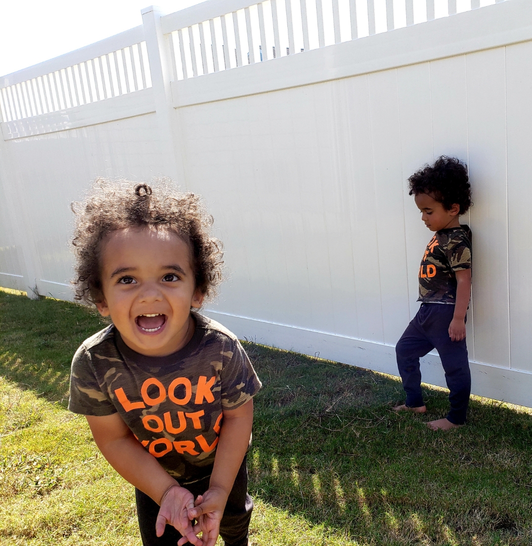 Twin boys playing in the backyard — one laughing at the camera, one exploring on his own